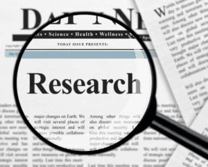 A magnifying glass zooms in on the word Research in bold print on a newspaper, emphasizing the focus on research-related news or topics. Text and headlines are partially visible in the background.