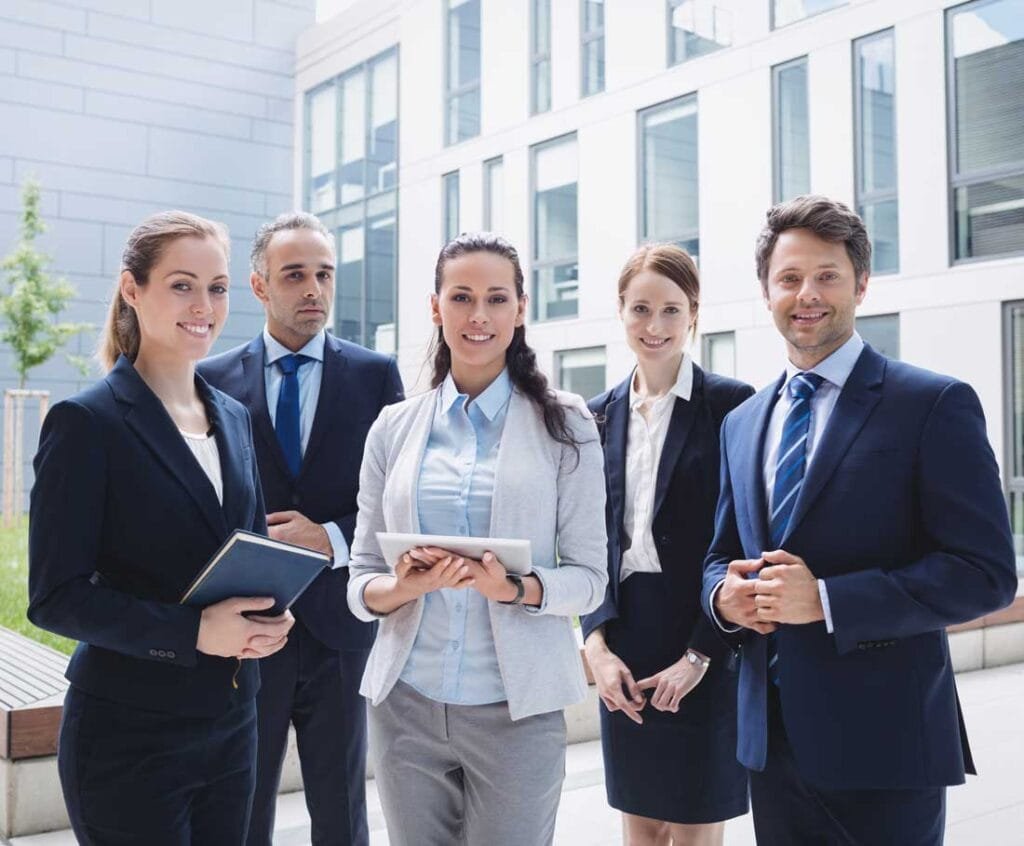 Five business professionals, three women and two men, stand outdoors in front of a modern office building. They are dressed in formal business attire and smiling, with two holding folders and one holding a tablet.
