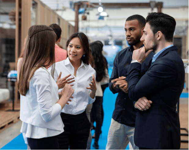 Four professionally dressed people stand in a group, engaged in conversation, inside a modern office or conference space with others in the background.