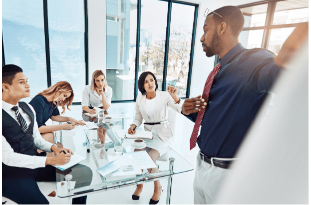 A man stands and presents to four colleagues seated at a glass conference table in a bright office. The group listens attentively, with laptops, documents, and coffee cups on the table.